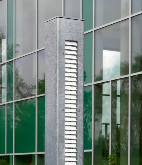 addo-l95-mini-mastleuchten-06 Nahaufnahme einer metallischen Struktur mit horizontalen Lamellen vor einem modernen Gebäude.