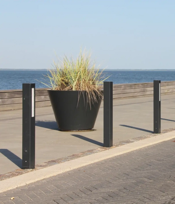 Ein großer schwarzer Pflanzkübel mit hohen, grasartigen Pflanzen, umgeben von drei schwarzen Pollerleuchten, in der Nähe eines Gewässers.