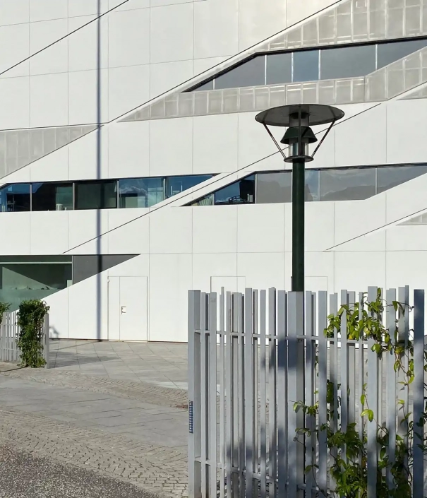 andersen-l207-mastleuchten-06 Straßenlaterne vor modernem Gebäude mit weißer Fassade und geometrischen Mustern.