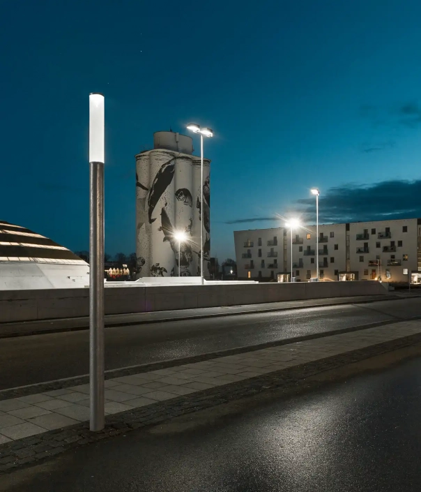 cover-l303-mastleuchten-12 Moderne urbane Szene bei Dämmerung mit einem großen, kuppelförmigen Gebäude und einer schlanken, zylinderförmigen Straßenlampe im Vordergrund.