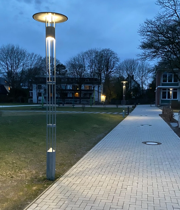 konus-l1012-mastleuchten-05 Gehweg in einem Park oder auf einem Campus bei Dämmerung, gesäumt von modernen Straßenlaternen und Bäumen, mit Gebäuden im Hintergrund.