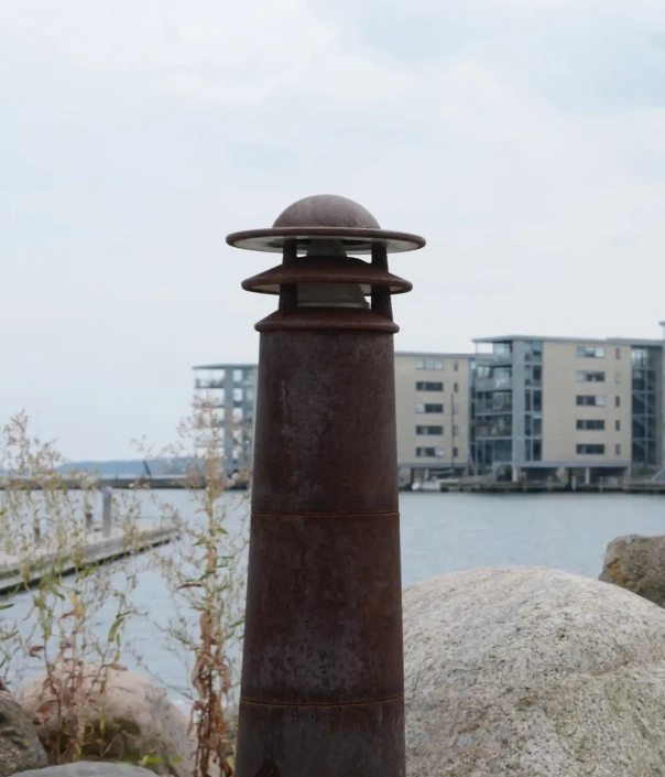 Eine ruhige Uferpromenade mit modernen Wohngebäuden im Hintergrund und einem verrosteten Metallpoller im Vordergrund.