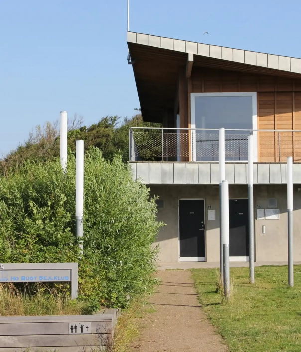 pin-l304-mastleuchten-07 Modernes Gebäude mit schrägem Dach und großen Fenstern, mit mehreren zylindrischen Lampen verschiedener Höhen im Vordergrund.