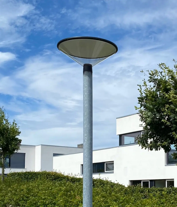 quintus-l500-mastleuchten-05 Moderne Straßenlaterne vor einem klaren blauen Himmel mit verstreuten Wolken und einem zeitgenössischen weißen Gebäude im Hintergrund.