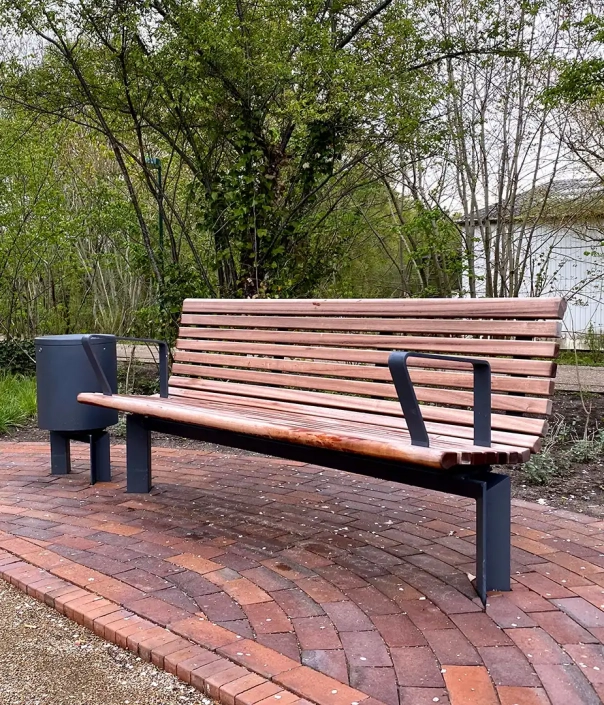 Parkbank mit Holzlatten und Metallgestell sowie einem integrierten Abfalleimer auf einem gepflasterten Weg.