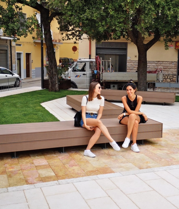 Zwei Personen sitzen auf einer modernen Holzbank in einem städtischen Außenbereich.