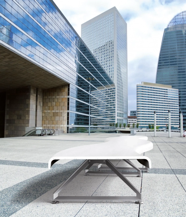 Eine moderne, weiße Bank mit Metallstützen in einem städtischen Umfeld mit hohen Glasgebäuden und einem großen offenen Platz.