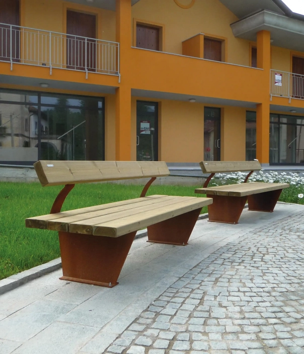 Zwei Holzbänke mit Metallstützen, die auf einer gepflasterten Fläche neben einem Gebäude platziert sind.