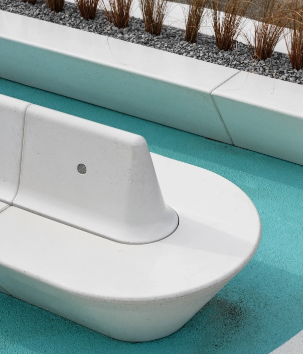 Moderne weiße Sitzbank aus Beton mit abgerundeten Formen in einem Außenbereich, umgeben von einem blauen Bodenbelag und dekorativem Kiesbett mit Pflanzen.