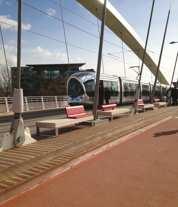 Moderne Sitzbänke aus Beton und Metall auf einer Fußgängerbrücke mit Straßenbahn und Stahlseilen im Hintergrund.