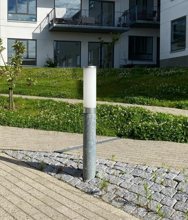 Eine moderne Außenleuchte in einem gut gepflegten Wohnbereich, die auf einer gepflasterten Fläche neben einem kleinen Baum steht.