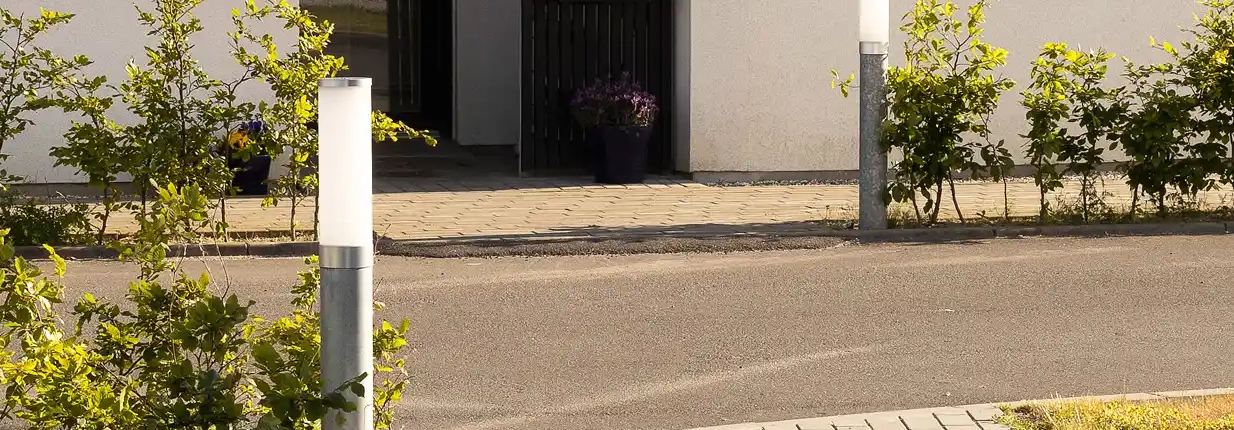 Das Bild zeigt den Eingang eines modernen Gebäudes mit einer schwarzen Tür und einem Glasfenster, flankiert von großen Fenstern. Vor dem Eingang befindet sich ein gepflasterter Weg mit grünen Sträuchern und zwei zylindrischen Außenleuchten.
