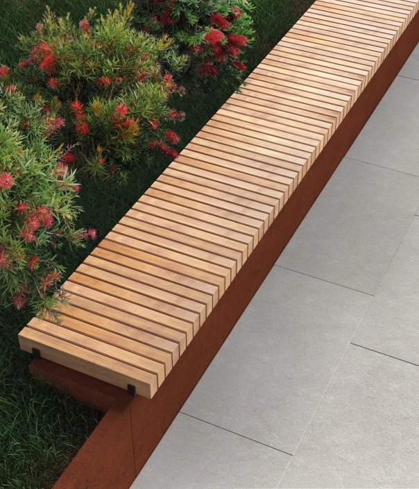 Nahaufnahme einer Bank mit Holzlatten und Cortenstahl-Untergestell, eingerahmt von Pflanzen.