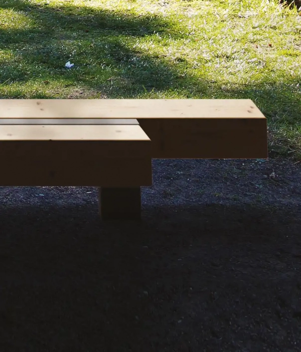 Holzbank mit minimalistischer, rechteckiger Form und zentraler Stütze, die die Sitzfläche in zwei Teile unterteilt. Die Bank steht auf einem naturbelassenen Boden im Freien.