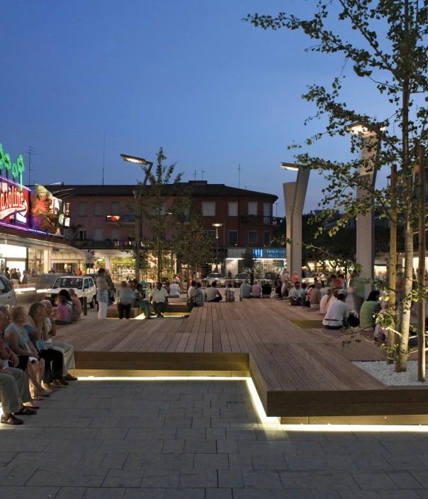Belebter Stadtplatz mit modularen Holzsitzbänken und stimmungsvoller LED-Beleuchtung bei Dämmerung.