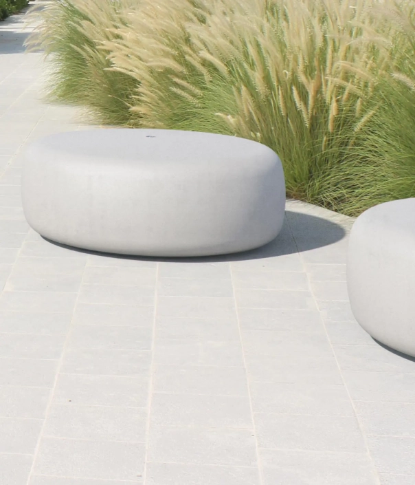 Zwei ovale, graue Steinhocker stehen auf einem hellen Pflasterweg, umgeben von grünem Schilfrohr.
