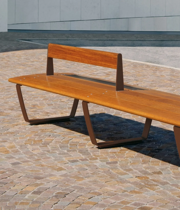 Eine moderne, Holzbank mit einer simplen, eleganten Form steht auf einem gepflasterten Boden. Im Hintergrund ist eine minimalistische Wand zu sehen.