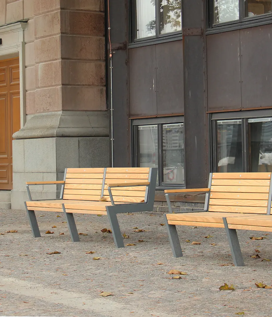 Zwei Holzbank-Bänke mit metallenen Gestellen stehen auf einem gepflasterten Boden, umgeben von herabgefallenen Blättern.