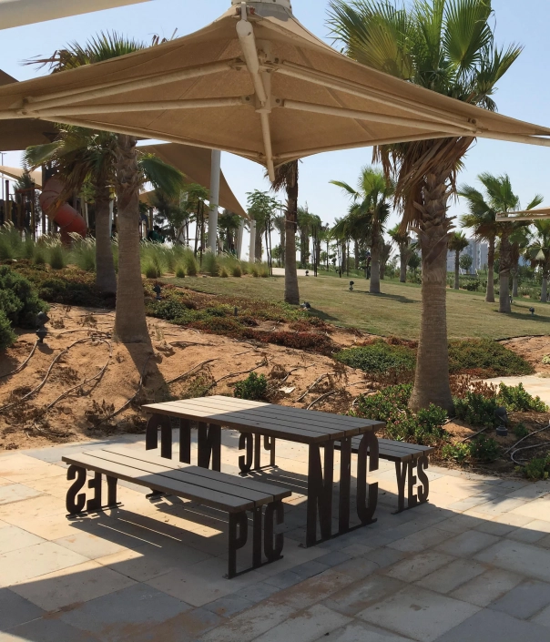 Zwei moderne Tisch-Bank-Kombinationen aus Holz und Metall stehen auf einem gepflasterten Gehweg in einem Garten, umgeben von Pflanzen und Sträuchern.