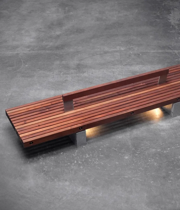 Eine moderne, stylische Bank aus Holz mit einem schlichten Design, auf einem grauen Betonboden platziert. Die Bank hat eine durchgehende Rückenlehne und ist mit integrierter Beleuchtung ausgestattet.