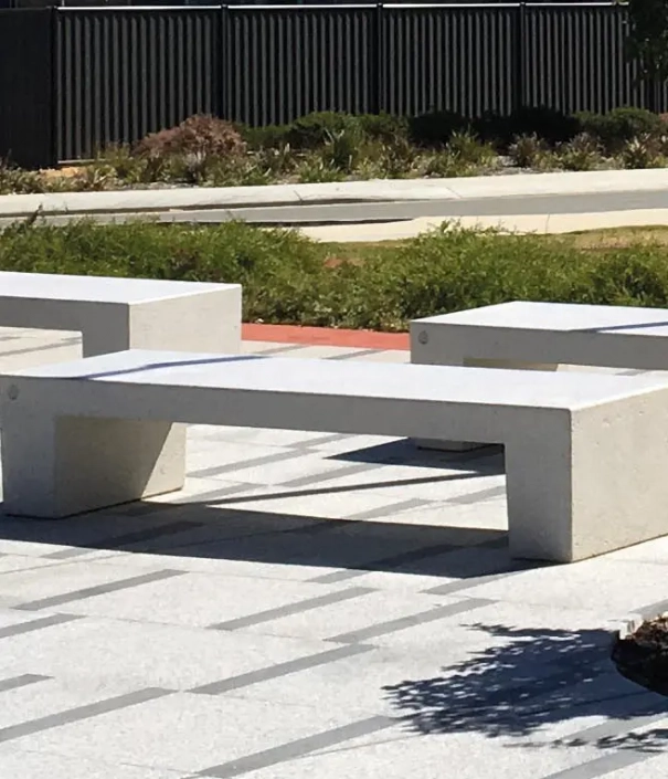 Ein moderner, grauer Betonblock steht in einem offenen, begrünten Außengelände. Umgeben von Rasen und Sträuchern sind mehrere ähnliche Blöcke auf einem gepflasterten Weg angeordnet.