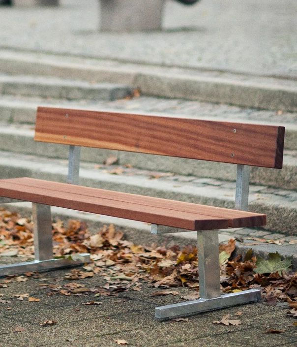 Eine leere Holzbank mit Metallgestell, umgeben von Laub auf dem Boden. Im Hintergrund sind Treppenstufen zu sehen.