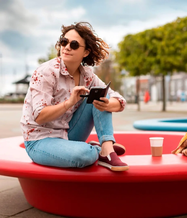 Junge Frau sitzt entspannt auf einer roten, runden Sitzbank im Freien und nutzt ihr Smartphone.