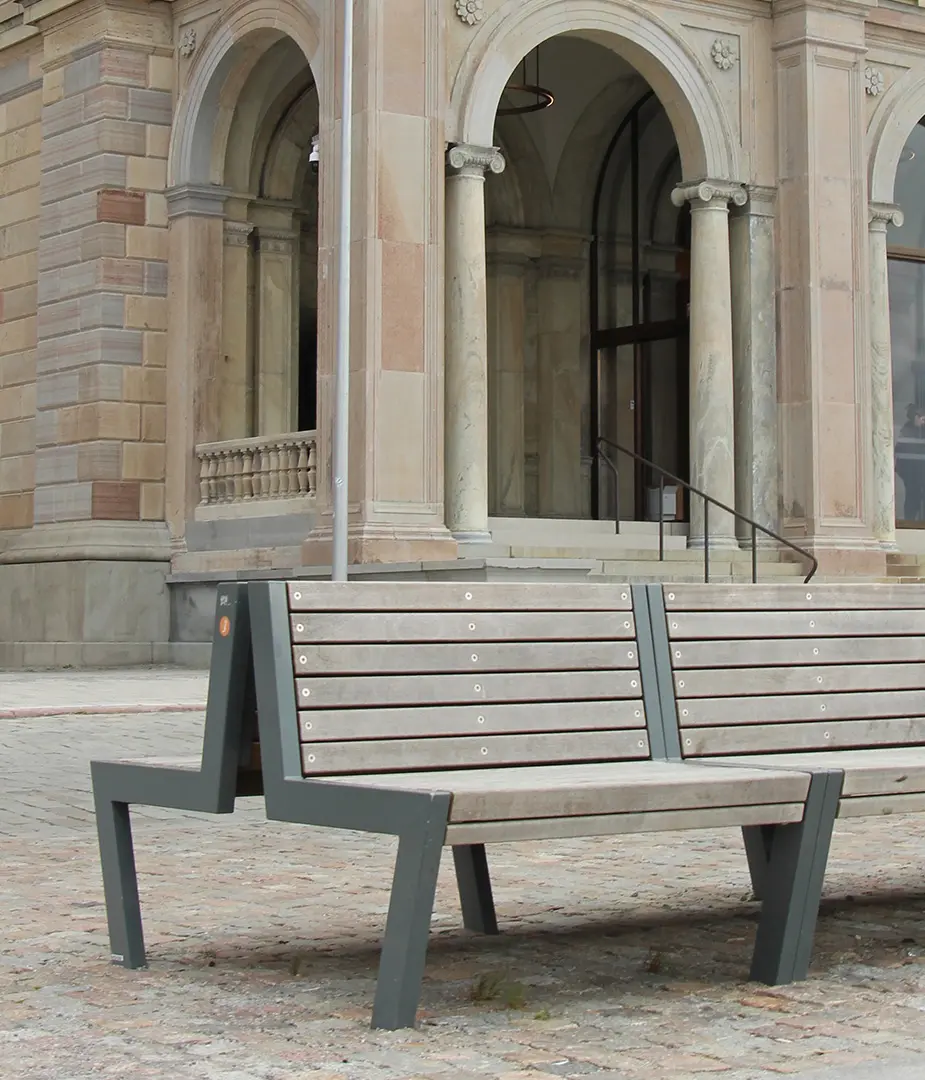 Blick auf eine moderne Bank aus Holz und Metall, die vor einem historischen Gebäude mit Säulen und großen Fenstern steht.