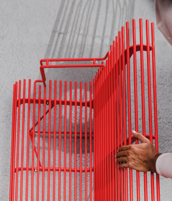 Hand einer Person berührt eine rote Drahtbank mit modularer Struktur und präzise gefertigten Metallstreben.