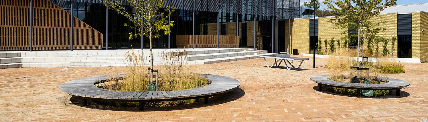 Runde Holzsitzbank um einen Baum in einem modernen, gepflasterten Außenbereich.
