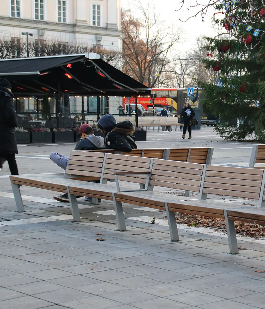 Zwei Personen sitzen auf einer Bank in einer städtischen Umgebung, während Passanten im Hintergrund vorbeigehen. Ein Weihnachtsbaum ist in der Nähe sichtbar.
