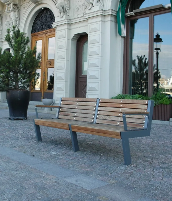 Eine moderne Holzbank mit Metallgestell steht auf einem gepflasterten Gehweg vor einem historischen Gebäude, umgeben von Pflanzen.