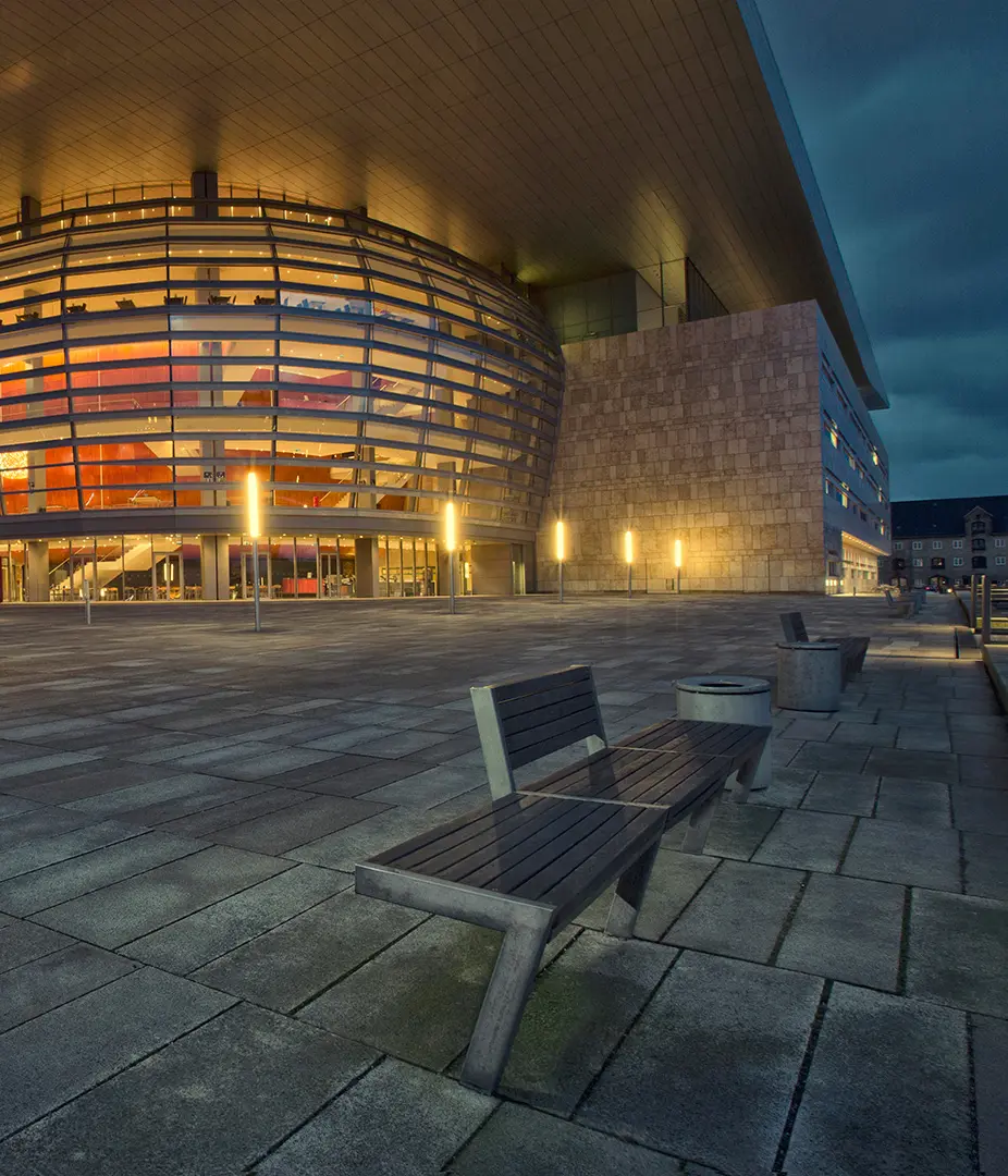 Exterioransicht eines modernen Gebäudes mit runder Fassade und großen Fenstern bei Dämmerung. Vor dem Gebäude befinden sich mehrere Bänke auf einem gepflasterten Platz.