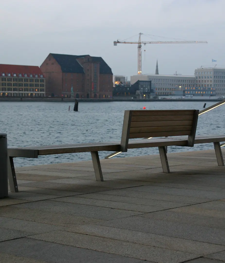 Eine ruhige Uferpromenade mit einer Holzbank im Vordergrund. Im Hintergrund ist das Wasser zu sehen, sowie moderne und historische Gebäude. Ein Kranausblick über das Wasser ist ebenfalls sichtbar. Der Himmel ist bewölkt und die Lichtverhältnisse sind düster.