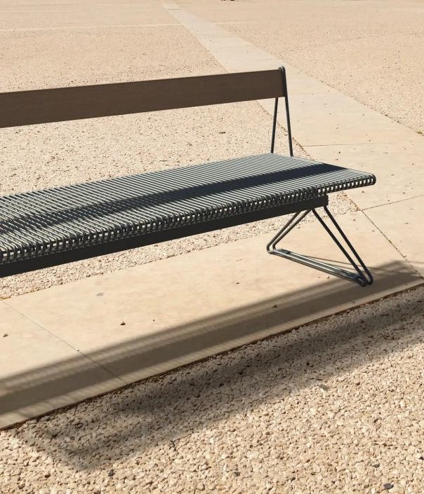 Eine moderne, graue Bank auf einer gepflasterten Fläche, umgeben von Kies. Die Bank hat ein minimalistisches Design mit einer abgeschrägten Lehne und einem stabilen, geschwungenen Gestell.