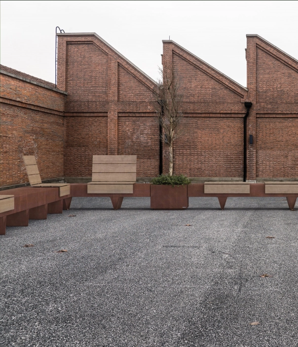 Eine moderne Sitzbank aus Holz und Metall, angeordnet in parallelen Reihen, steht vor einer Backsteinwand. Um die Bank herum sind kleine Gräser oder Pflanzen angeordnet. Der Boden ist mit Kies bedeckt.