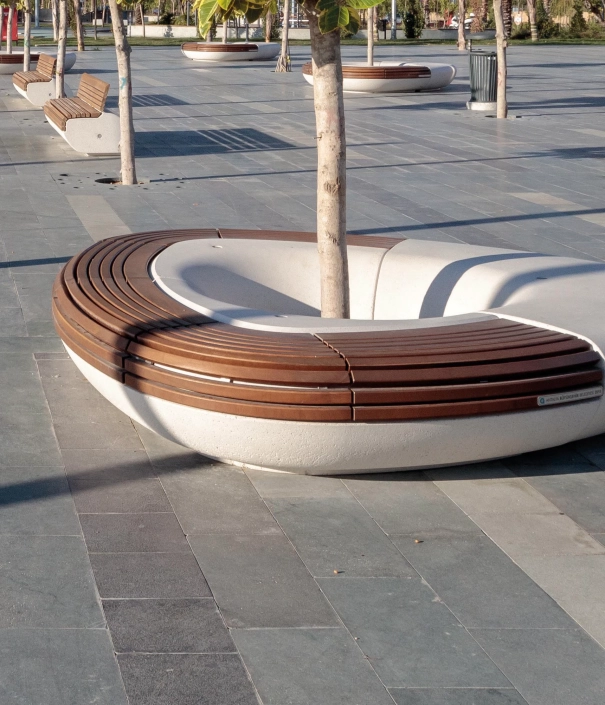 Öffentlicher Platz mit kreisförmigen Sitzbänken um Bäume, umgeben von einer modernen Stadtlandschaft.