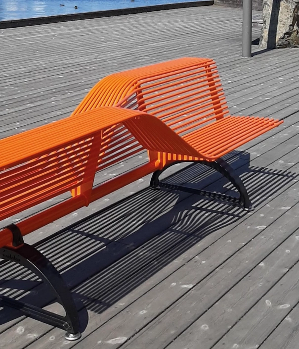 Orangefarbene Metallbank mit geschwungener Rückenlehne auf einer Holzplattform nahe dem Wasser.