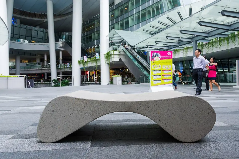 Ein elegantes, modernes Bankelement aus hellem Beton, das in einem offenen, urbanen Bereich steht. Im Hintergrund sind Menschen zu sehen, die im Gebäude herumgehen, sowie eine Stele mit Informationen. Die Umgebung wirkt zeitgenössisch und architektonisch ansprechend.