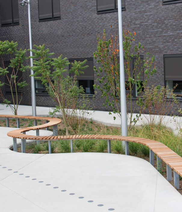 Alt-Text: Eine Ansichtsaufnahme eines modernen Außenbereichs mit geschwungenen Holzweg und bepflanzten Beeten. Im Hintergrund sind moderne Gebäude mit großen Fenstern zu sehen.