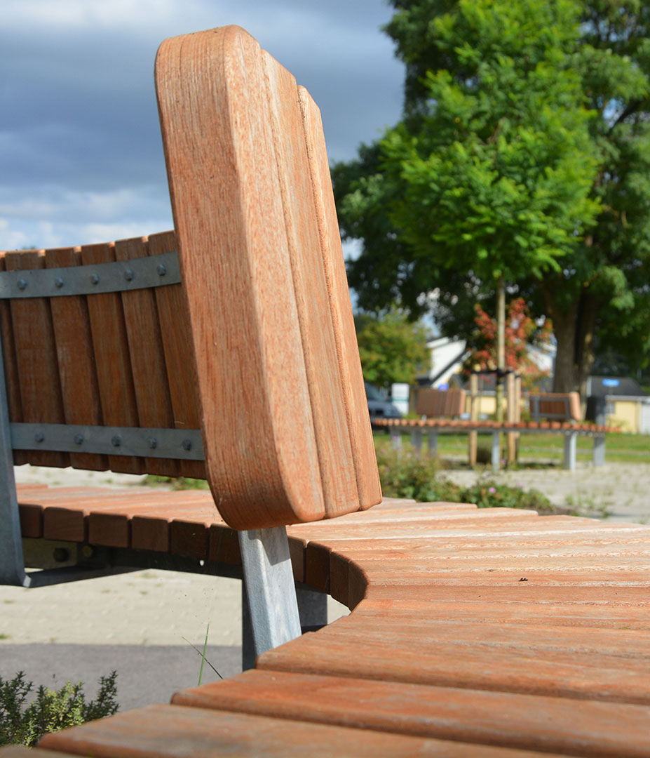 05Maja_Hitsa_Seite Detailaufnahme einer modernen Holzbank mit einem metallischen Gestell, im Hintergrund sind Bäume und weitere Sitzgelegenheiten sichtbar.