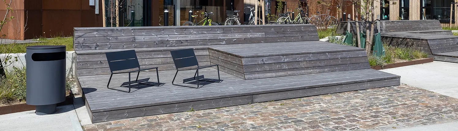 Hölzerne Sitzgelegenheit mit zwei schwarzen Stühlen und einem kleinen Tisch, umgeben von neu gestalteter urbaner Landschaft. Im Hintergrund sind Fahrräder und Gebäudefassaden zu sehen.