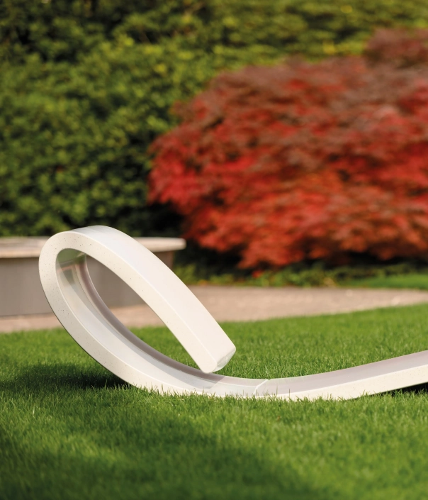 ine moderne, geschwungene Liege aus hellem Material liegt auf grünem Rasen, umgeben von buntem Laub im Hintergrund.