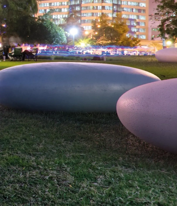 Zwei große, ovale Skulpturen auf einer grünen Wiese, umgeben von schwachem Licht bei Nacht. Im Hintergrund sind Lichtreflexe von Gebäuden und Menschen sichtbar.