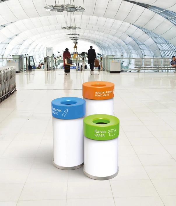 Drei Abfallbehälter in verschiedenen Farben (Orange, Grün, Blau), beschriftet mit "Gemischter Müll", "Papier" und "Kunststoff", stehen in einer modernen, hellen Bahnhofshalle.