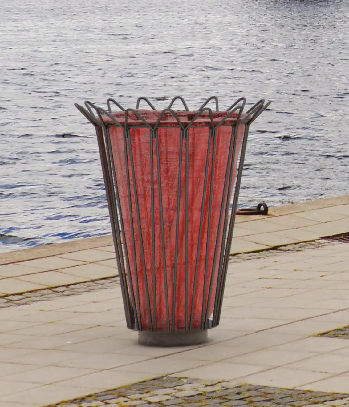 Ein moderner Mülleimer mit einer roten, gitterartigen Struktur steht an einem Uferweg neben einem Gewässer.
