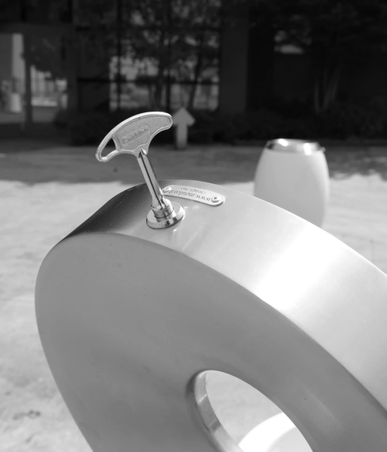 Ein metallisches Objekt in Form der Zahl „0“ mit einem Aufziehschlüssel an der Oberseite, in einer urbanen Umgebung. Im Hintergrund sind unscharf weitere ähnliche Objekte und Pflanzen zu erkennen.