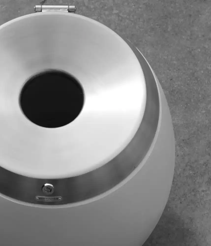 Eine runde, moderne Mülltonne mit einer metallenen Oberseite und einem runden Einwurfloch, auf einem grauen Boden.