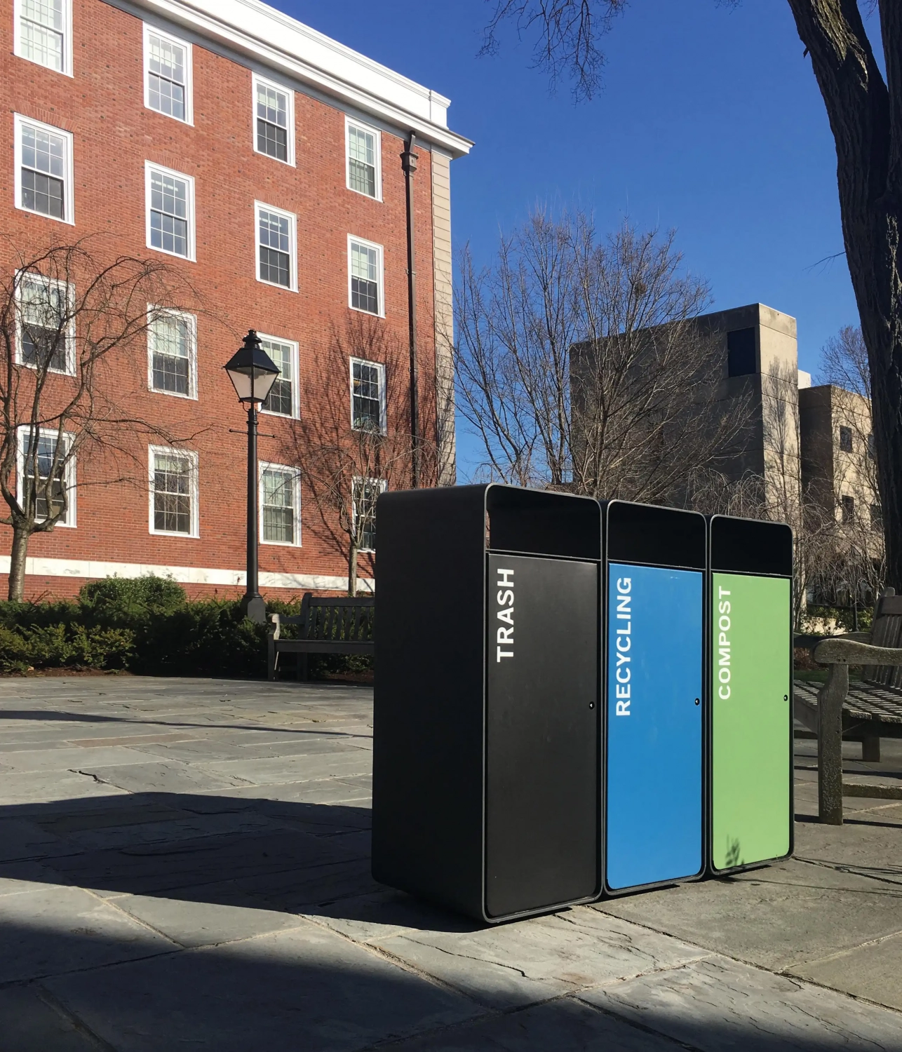 box-solid-abfallbehaelter-05 Drei Abfallbehälter in einem Park, beschriftet mit "TRASH", "RECYCLING" und "COMPOST", vor einem Hintergrund von Gebäuden und Bäumen.