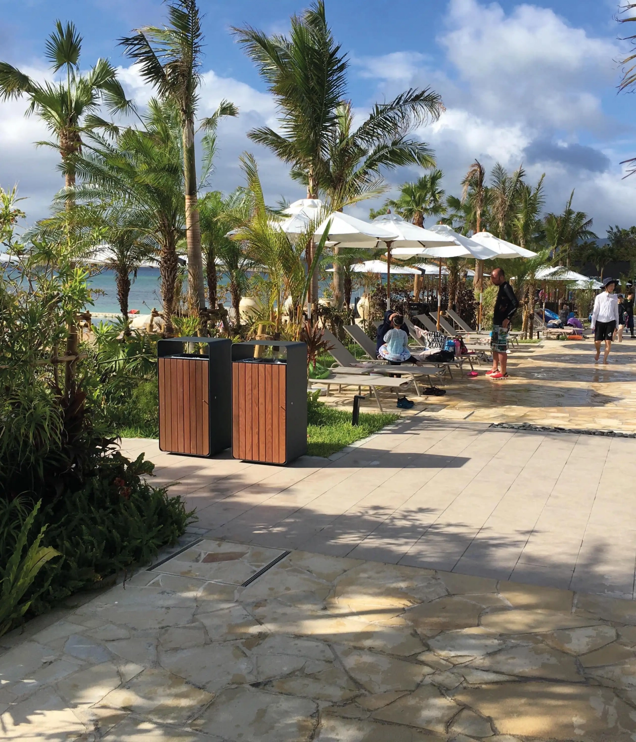 Eine Aussicht auf eine Strandpromenade mit Liegen und Sonnenschirmen, umgeben von Palmen. Im Vordergrund stehen zwei Mülleimer, während im Hintergrund Menschen entspannen und sich am Strand aufhalten.